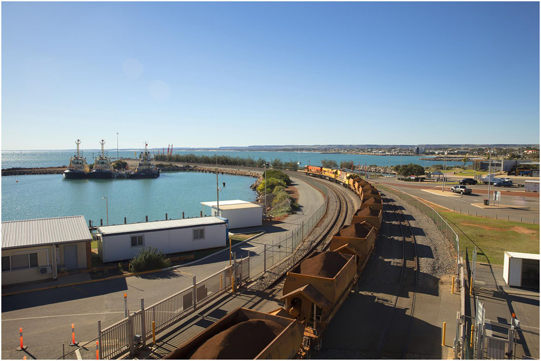 View of loaded train cars curved along groyne with 3 tugboats on left and parkland on right. Ocean in background.